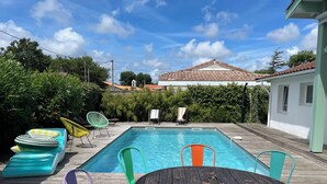 Una piscina rectangular con una cubierta de madera, rodeada de sillas coloridas y una mesa de madera redonda.