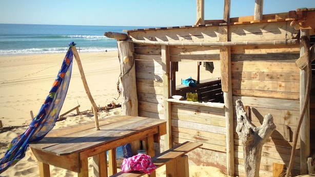 Una cabaña de playa de madera con una banca y una hamaca, con vista al océano.