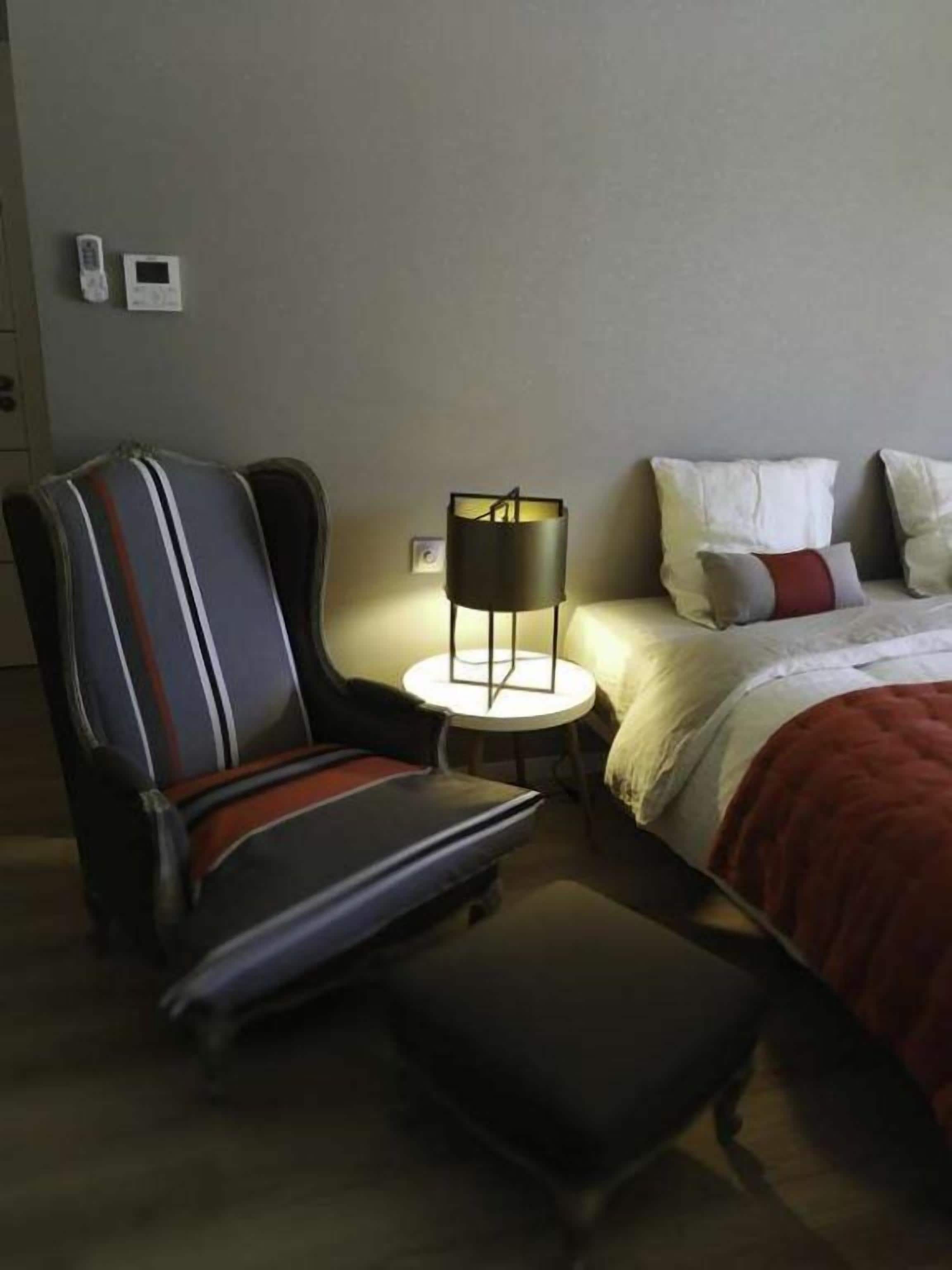 A hotel room with a bed, a chair, a small table, and a lamp.