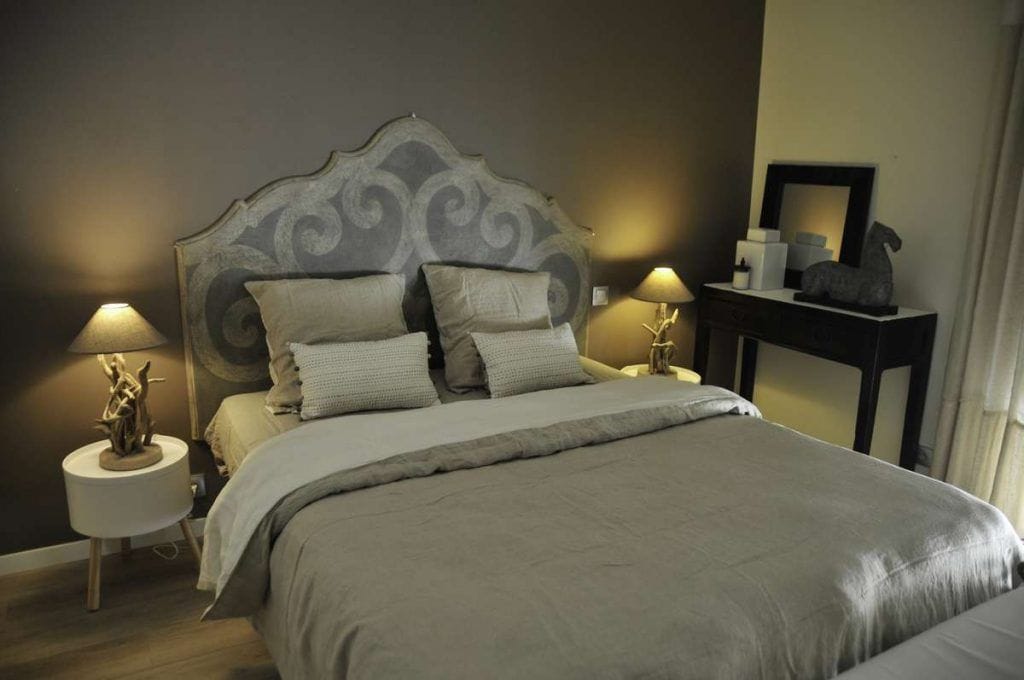 Una cama bien hecha con cabecera gris, dos lámparas de noche y una mesita con una estatua y un espejo.