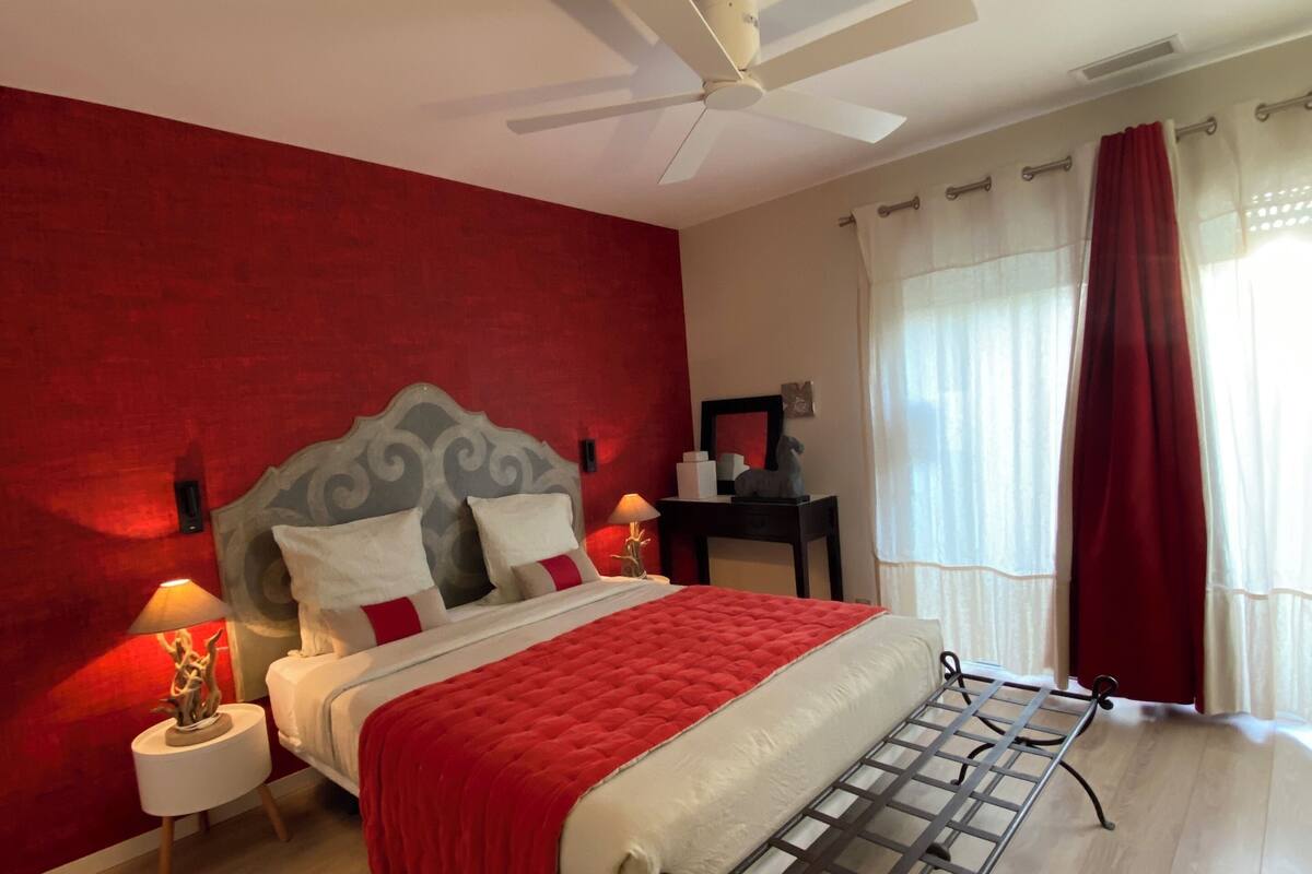 Une chambre à coucher avec un lit, un ventilateur de plafond, une table de chevet avec une lampe, une fenêtre avec des rideaux et un plancher en bois.