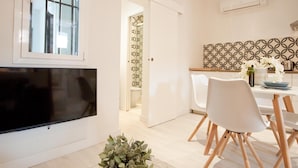 A modern living room with a flat-screen TV, a dining area with white chairs and a table, and a kitchen with patterned backsplash and white cabinets. - A perfect refuge in Madrid's city center (Madrid)
