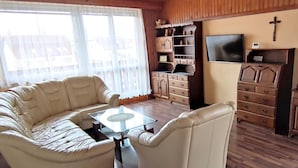 A living room with a sofa, armchair, glass coffee table, wooden cabinet, TV, and a cross on the wall.