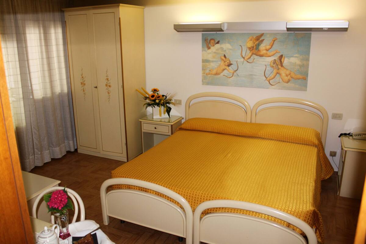 A bedroom with a double bed, a nightstand, a bedside table with a lamp, a wardrobe, and a painting of cherubs on the wall.