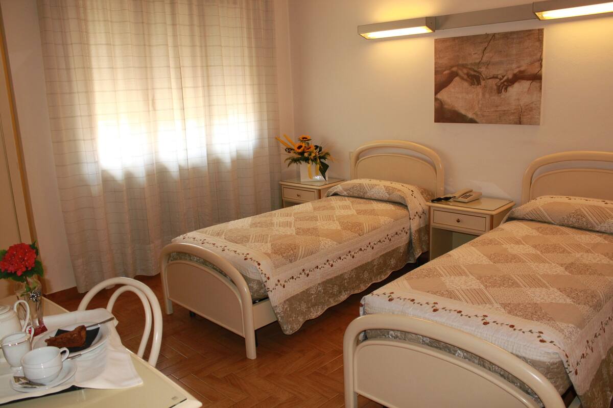 A bedroom with two beds, a bedside table with a lamp, a small table with cups and a vase of flowers, and a painting on the wall.