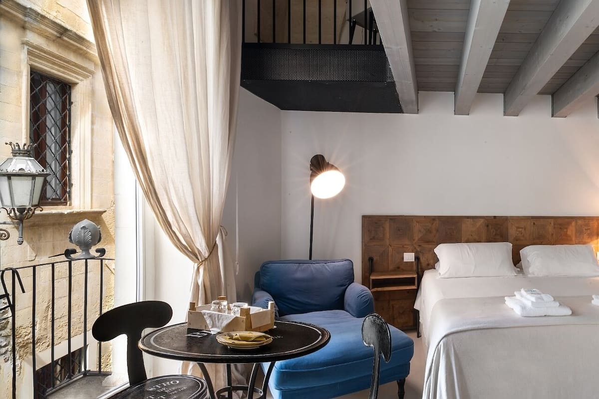 A hotel room with a bed, a blue chair, a small table, a lamp, and a staircase.