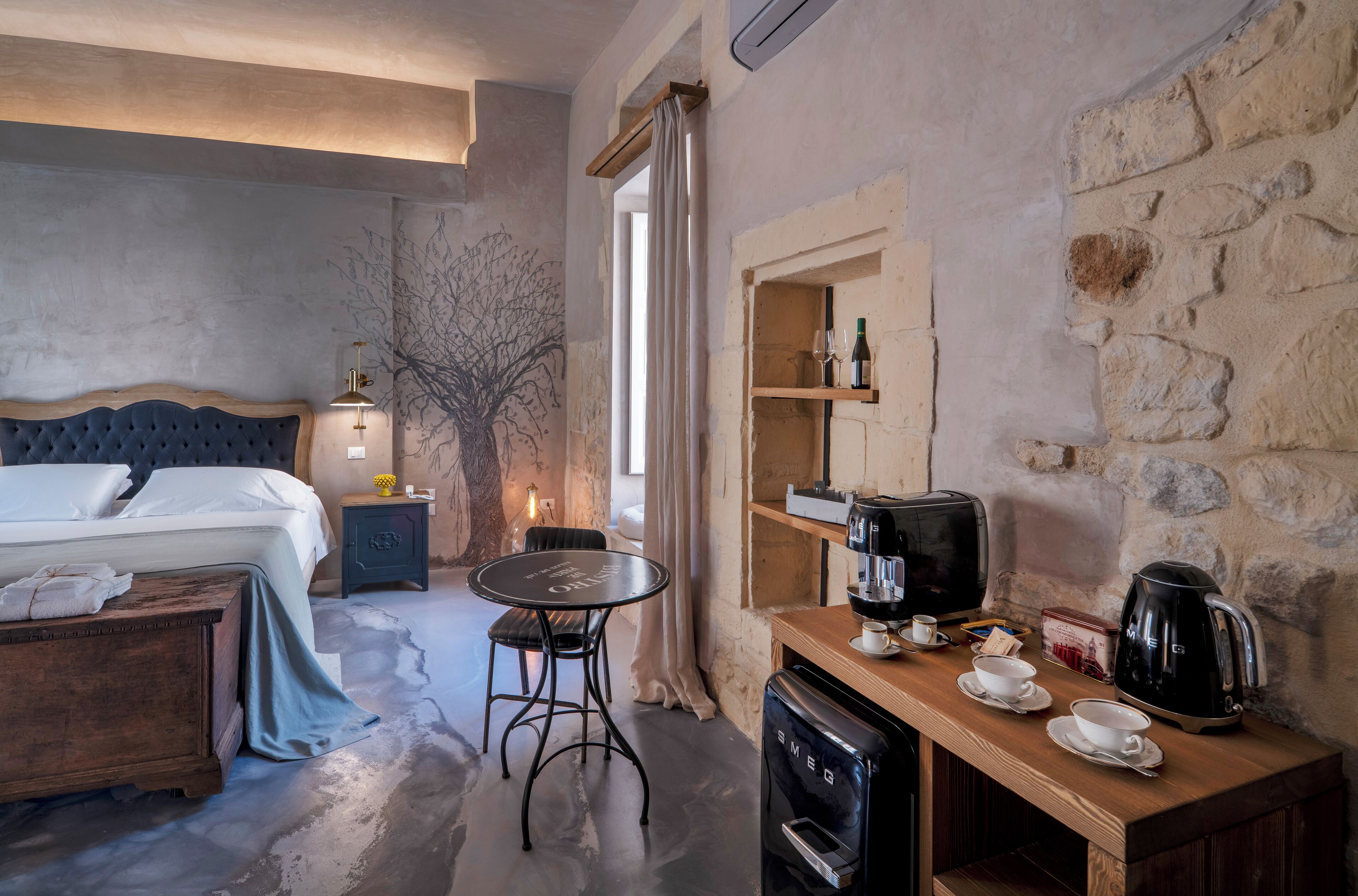 A bedroom with a bed, bedside table, and a small desk with a coffee maker and cups.