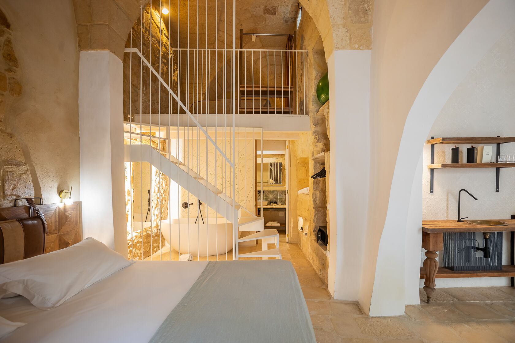 Un dormitorio con una cama grande, una escalera blanca, una pared de piedra y una zona de cocina con estantes de madera.