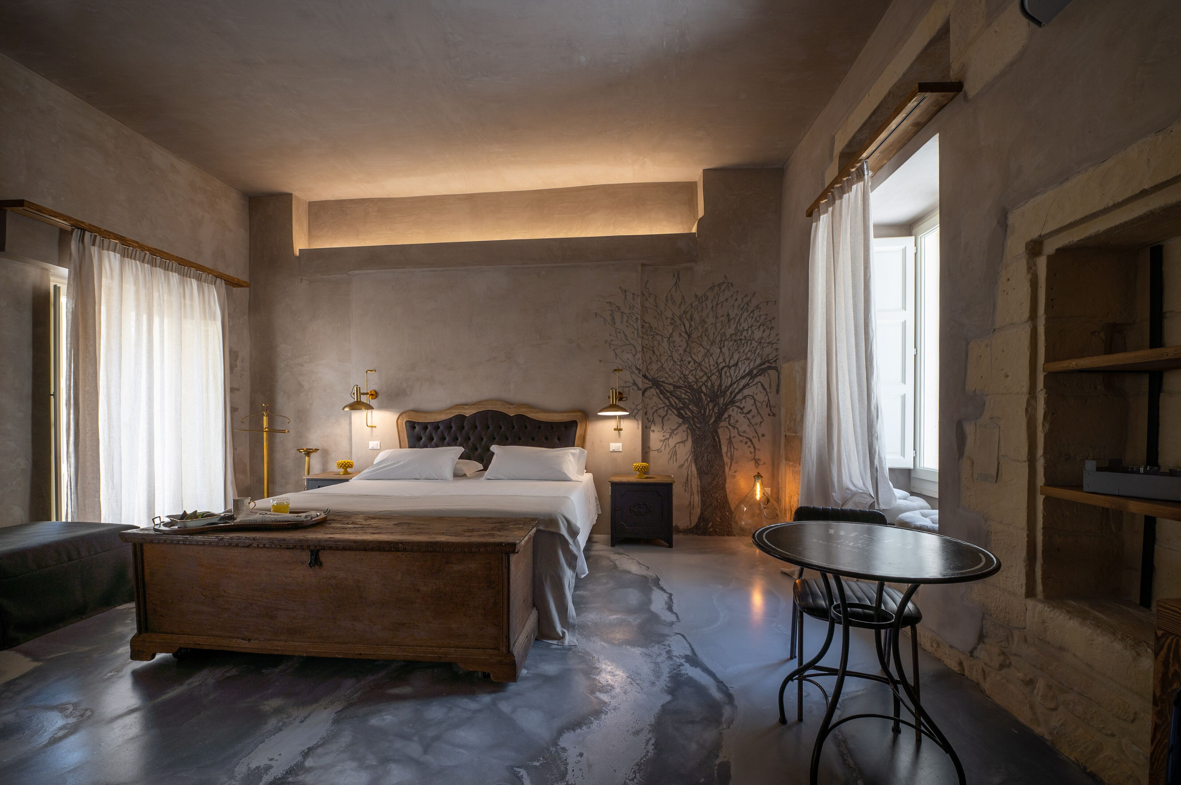 Un dormitorio con una cama grande, mesitas de noche, un cofre de madera, una mesa redonda y un mural con motivos de árboles.