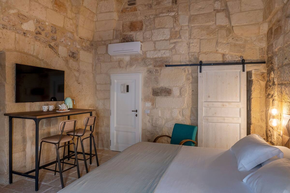 A room with a stone wall, a bed with white bedding, a desk with two chairs, a television mounted on the wall, and a door with a handle.