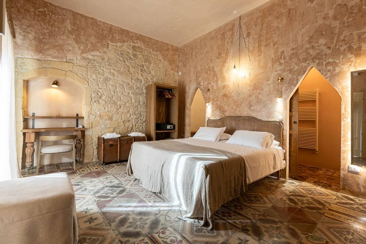 A bedroom with a stone wall, a bed with white linens, a wooden desk, and a patterned floor.