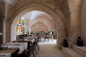 Un comedor de paredes de piedra con mesas y sillas, una ventana de vitral y un muro de ladrillo al fondo.