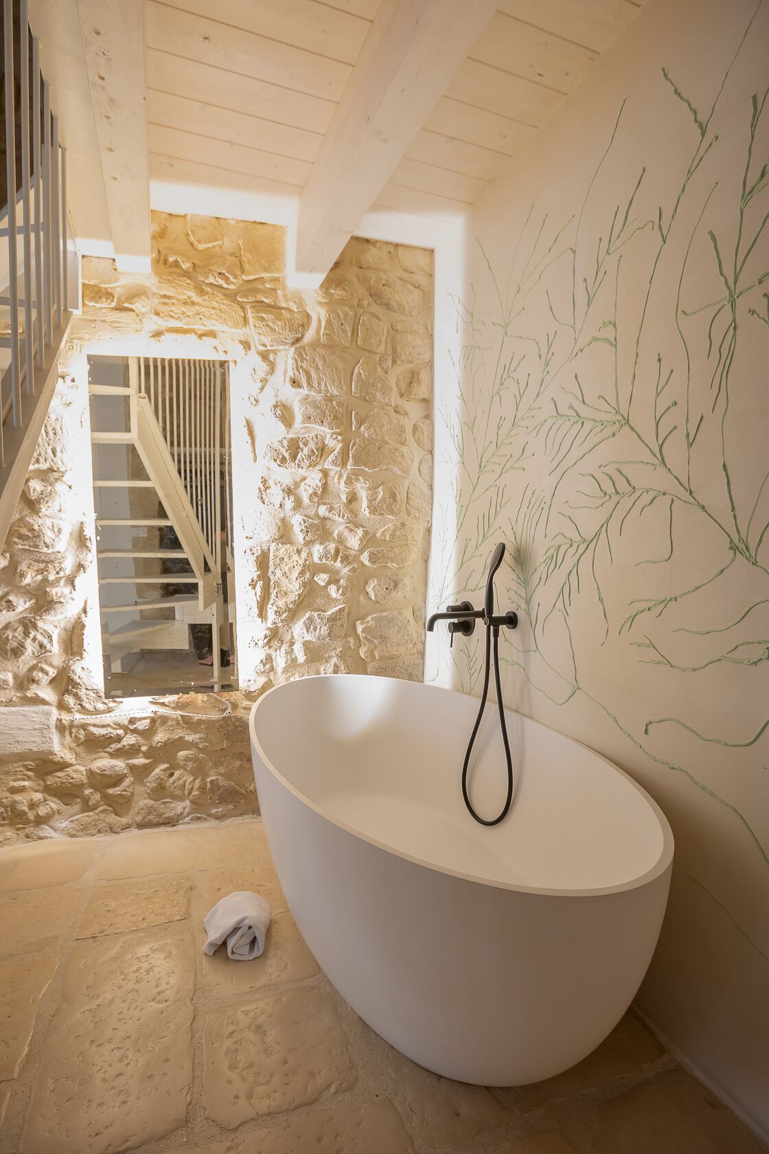 Un baño moderno con una bañera independiente, paredes de piedra y una escalera que conduce a un nivel superior.