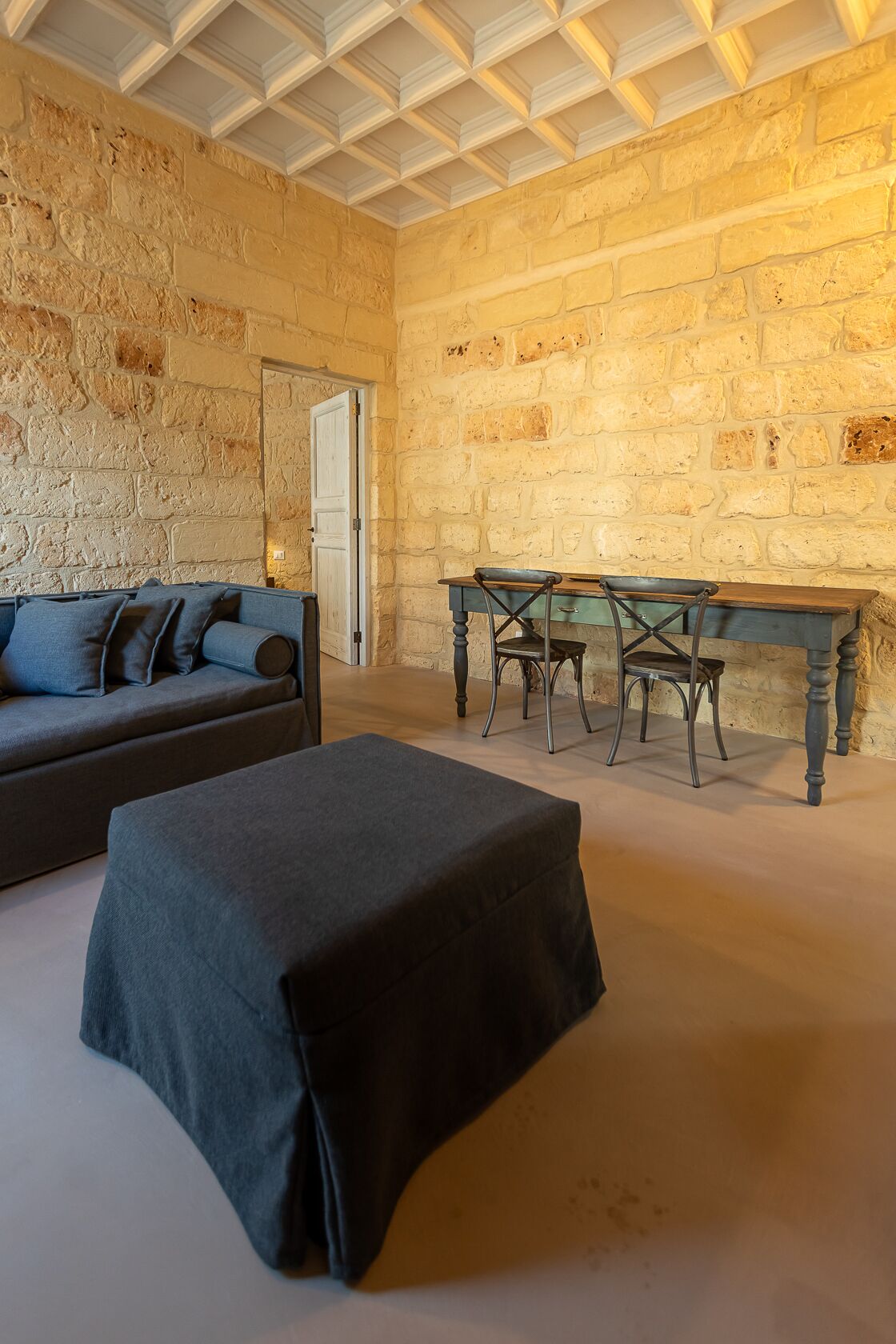 Ein Zimmer mit Steinwand, einem Holztisch mit Stühlen, einer Couch und einem abgedeckten Hocker.