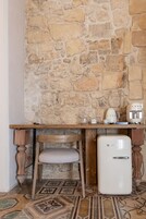 A rustic kitchen with a stone wall, a wooden table, a vintage white refrigerator, and a chair.
