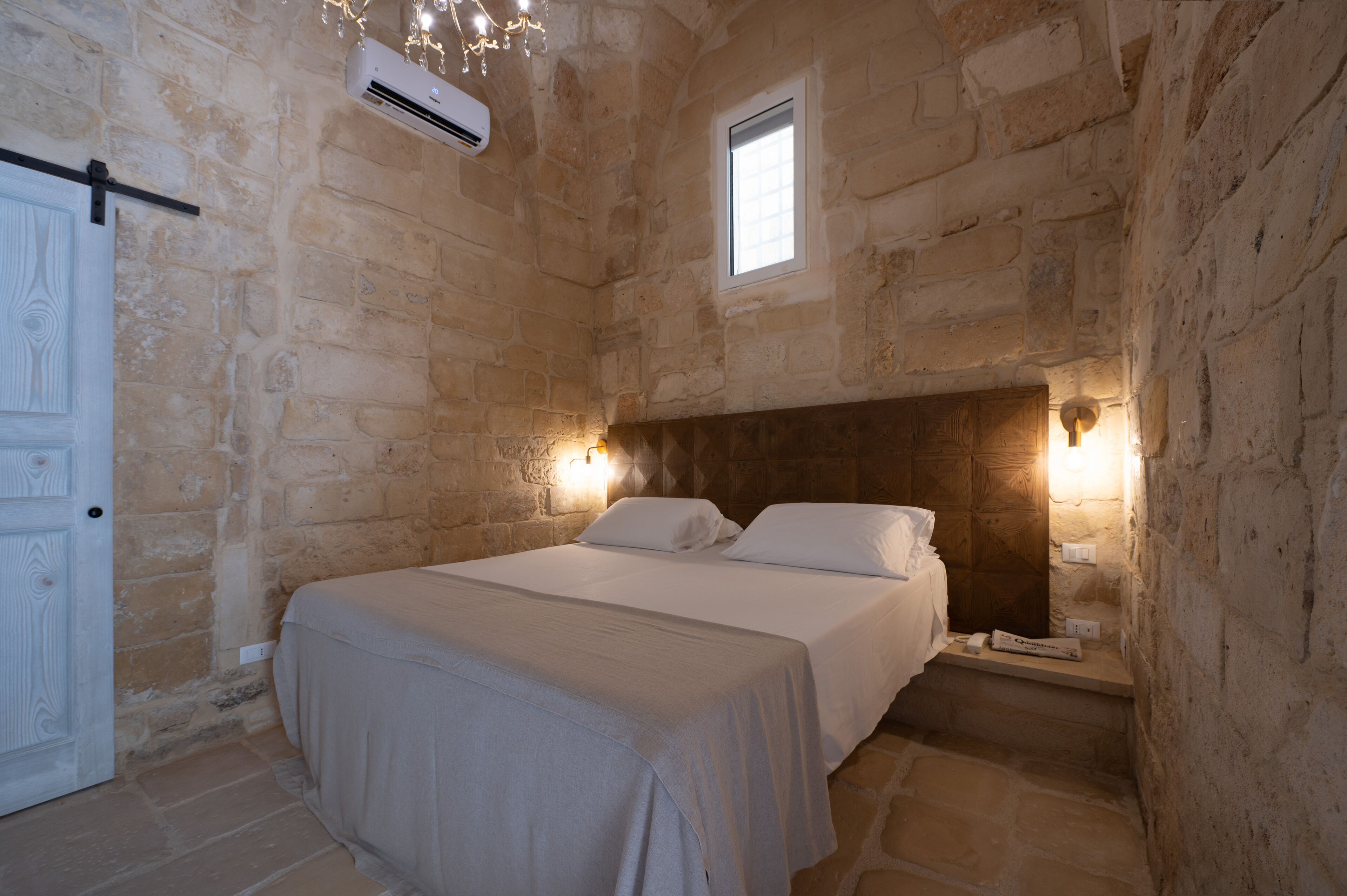 Dormitorio con paredes de piedra, una cama, un ventanal y un aire acondicionado instalado en la pared.