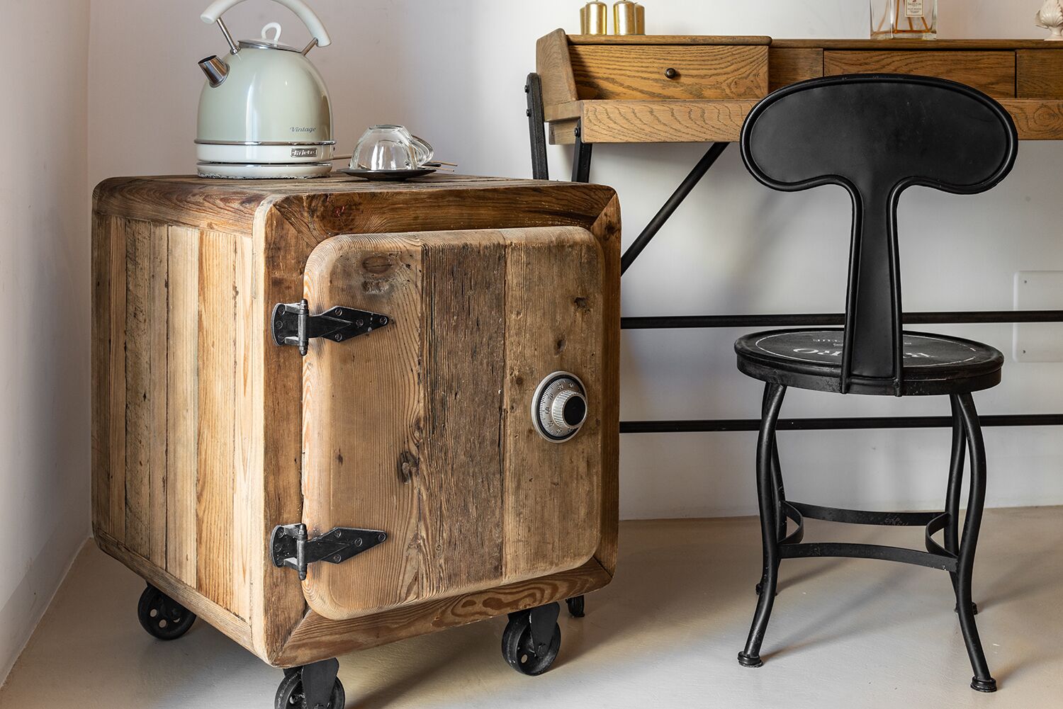 A wooden cabinet with wheels, a vintage kettle, and a black chair.