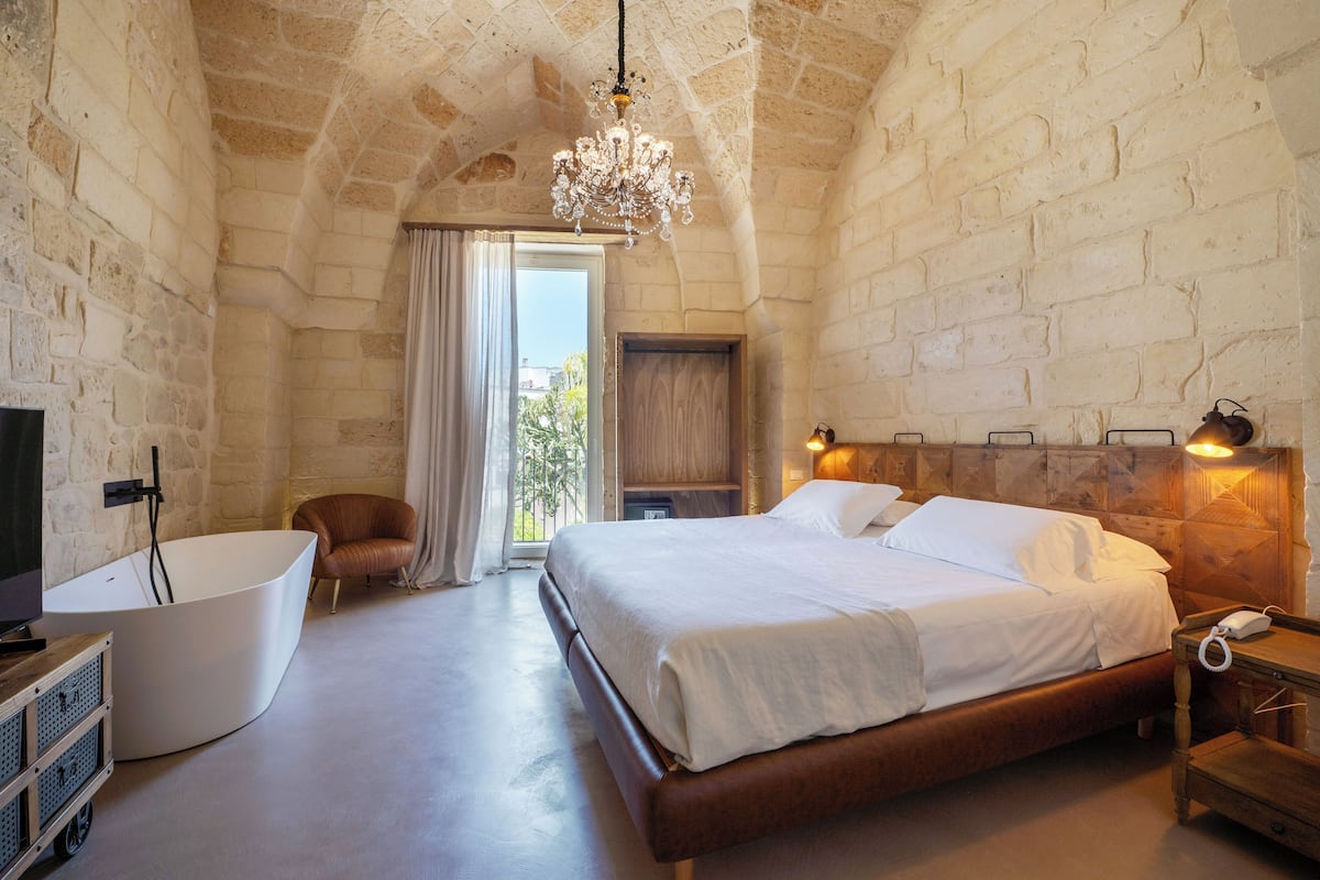 A bedroom with a large bed, a bathtub, a wooden nightstand, and a chandelier.