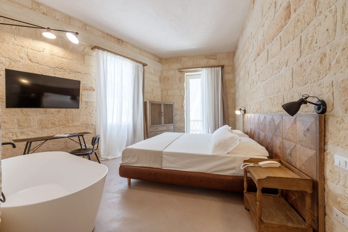 A bedroom with a stone wall, a bed, a bedside table, a lamp, a TV, and a window with curtains.