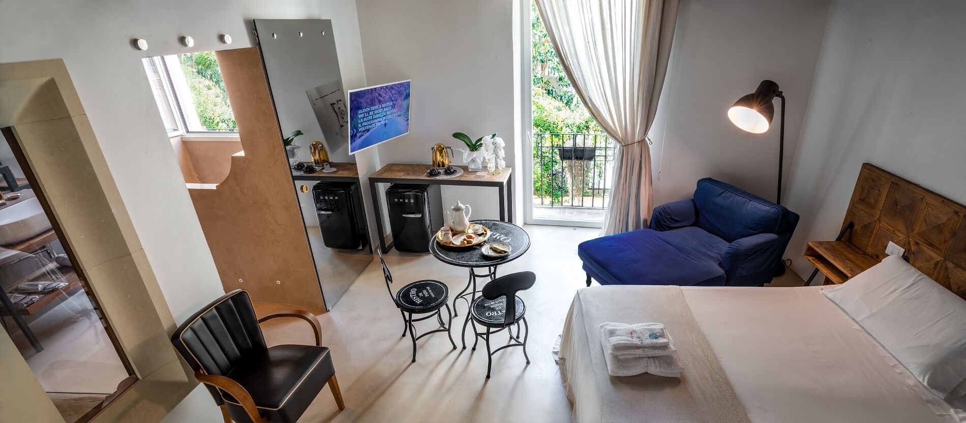 Habitación de hotel pequeña con cama, una mesa de comedor con sillas, televisor y balcón con cortinas.