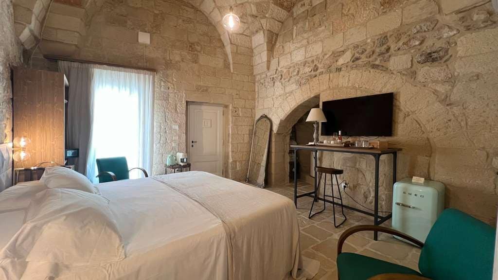 A hotel room with a stone wall, a bed, a desk, a chair, and a television.