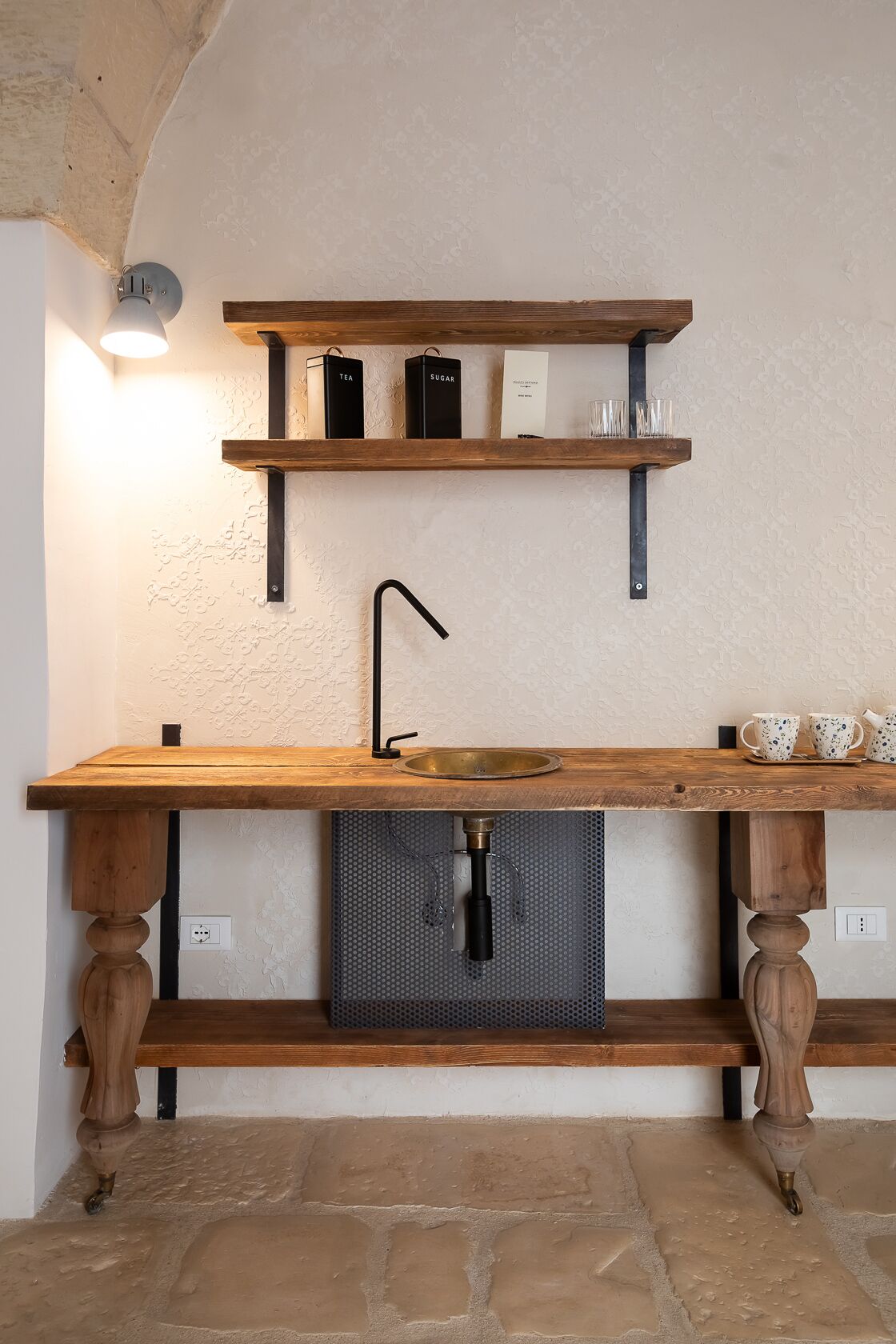 Eine rustikale Küche mit einer Holzauslage, einem Spülbecken und Regalen darüber, auf denen Teebohnen und Zuckerbehälter stehen.