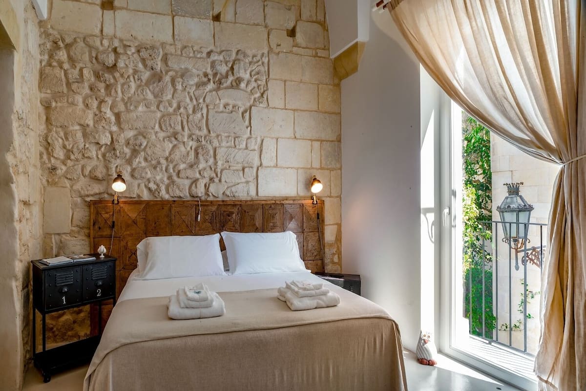 A bedroom with a stone wall, a wooden headboard, a bed with white linens, two bedside lamps, and a window with curtains.