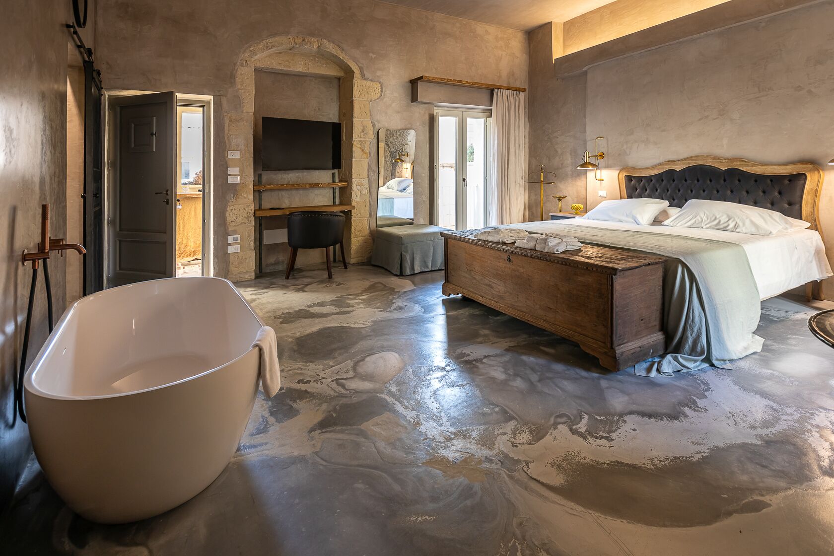 A spacious bedroom with a large bathtub, a wooden bench, a bed with white linens, and a stone wall.
