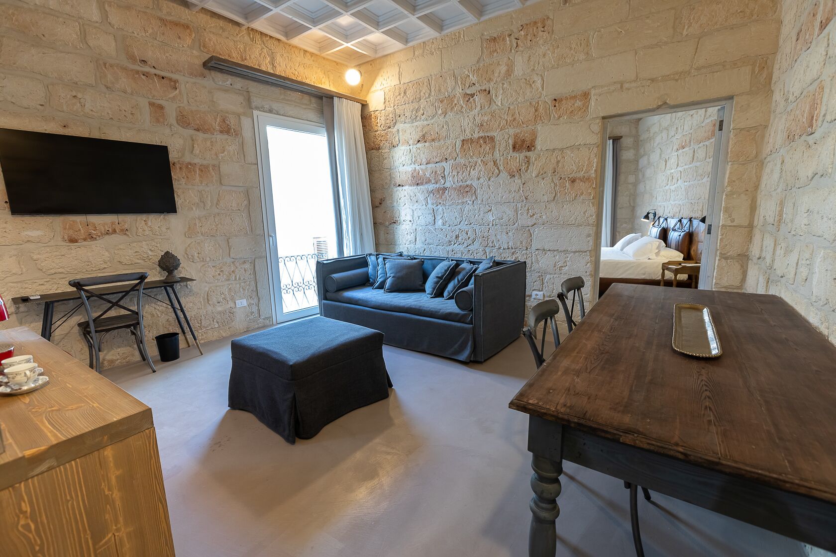 A cozy room with a sofa, a wooden table, a TV mounted on the wall, and a balcony with a view.