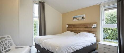Un dormitorio con cabecera de madera, una cama individual, una silla blanca, una mesita de noche pequeña y una ventana con cortinas.