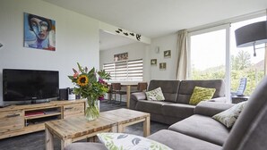 A modern living room with a flat-screen TV, a wooden coffee table, a grey sofa, and a large window offering a view of the outdoors.