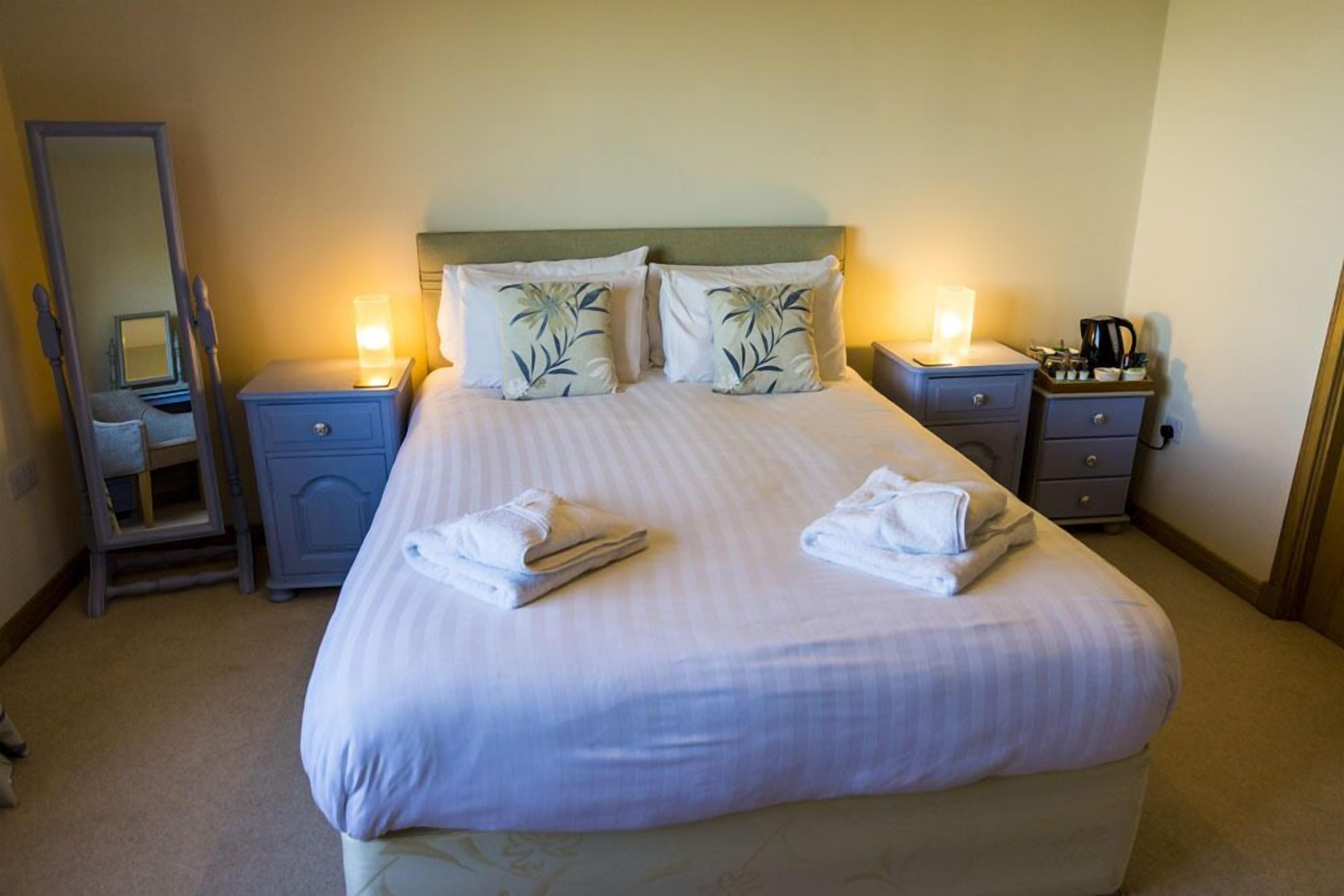 A neatly made bed with pillows and folded towels, a bedside table with a lamp, a mirror, and a small cabinet.