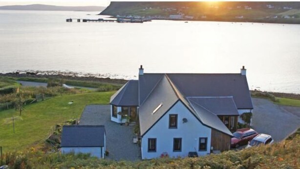 A house with a sloped roof, a small porch, and a driveway with parked cars. - Old Pier Cottage B&B (Uig)