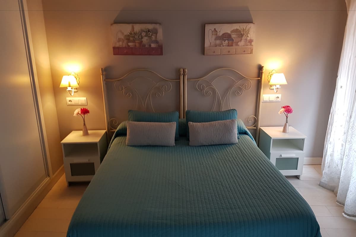 A neatly made bed with a teal bedspread and two pillows, flanked by two bedside tables with lamps and small vases of flowers.