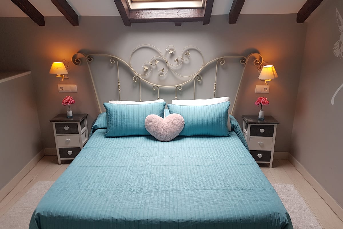A bedroom with a skylight, a bed with teal bedding, heart-shaped pillows, and bedside tables with lamps and vases.