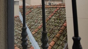 Vista a través de una ventana con barandilla de metal, que muestra un puente de piedra con arco, un tejado de tejas y un veleta.