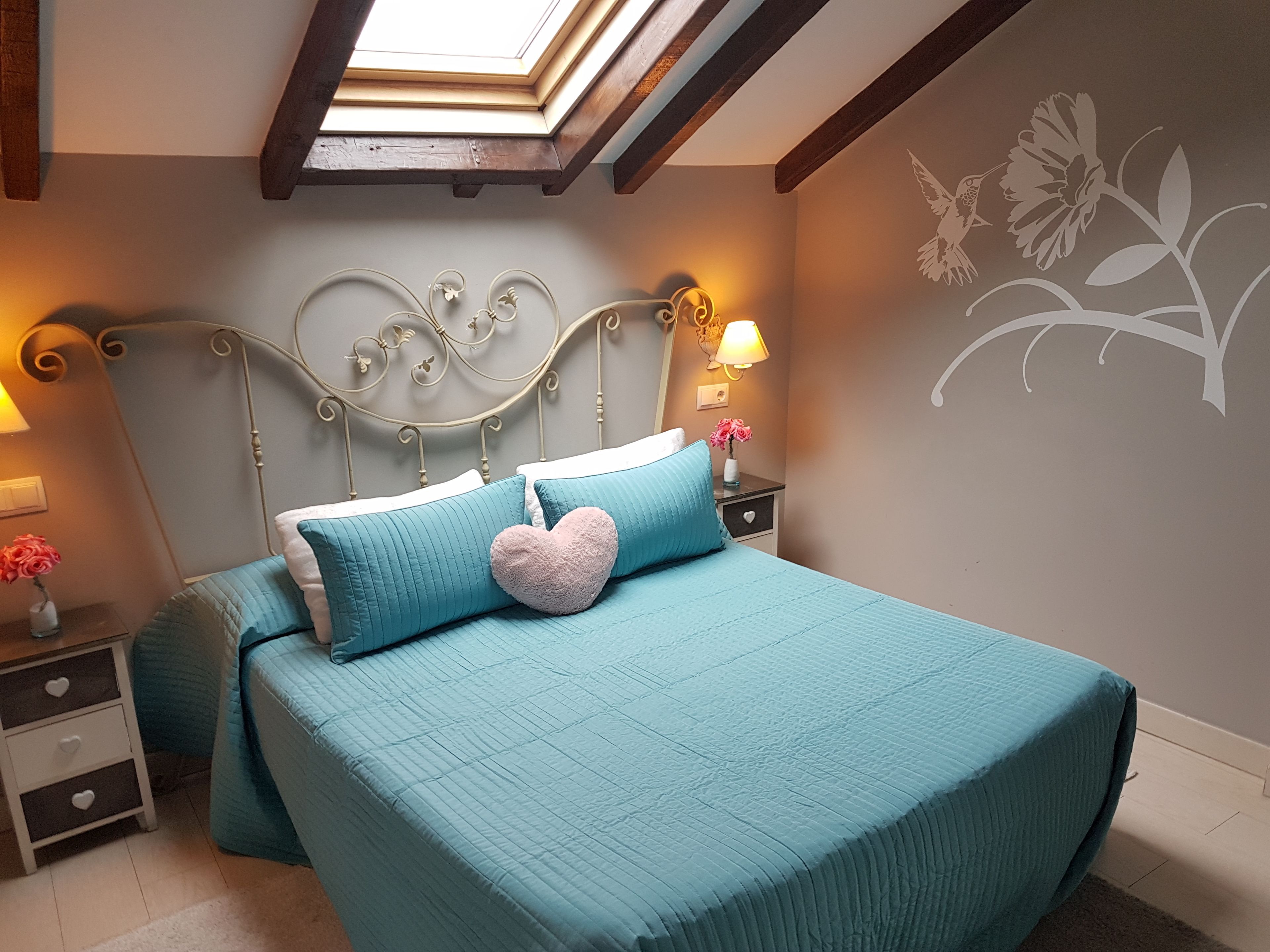 A bedroom with a skylight, a bed with teal bedding, a heart-shaped pillow, and a wall mural.