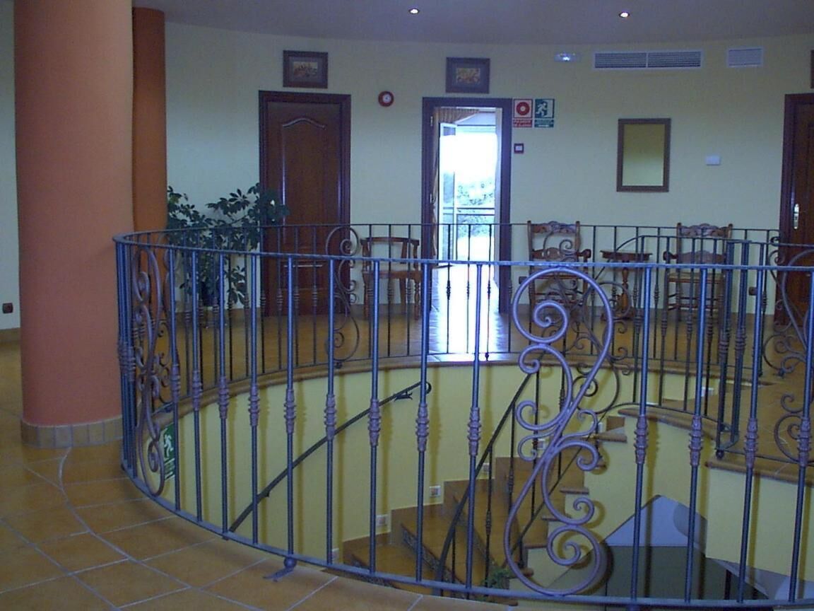 Un área interior amplia con barandilla metálica curvada, sillas de madera y una escalera que sube a un nivel superior.