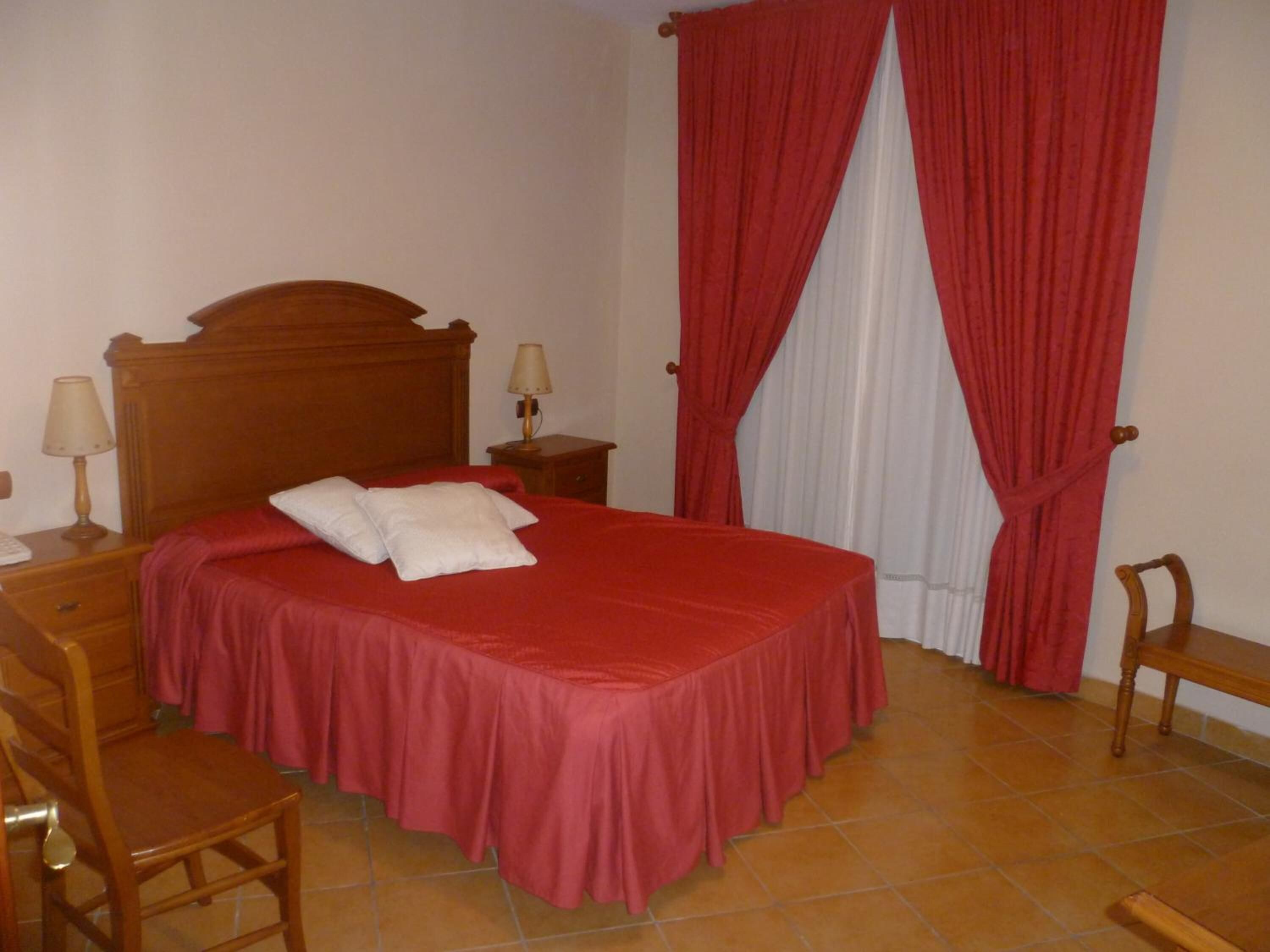 A neatly made bed with a red bedspread and two pillows, a wooden nightstand with a lamp, a wooden chair, and a window with red curtains.