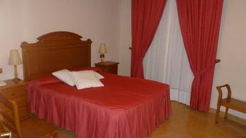 A neatly made bed with a red bedspread and two pillows, a wooden nightstand with a lamp, a wooden chair, and a window with red curtains.