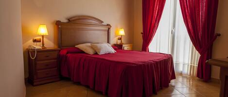 A bedroom with a wooden headboard, two bedside lamps, a telephone, a red bedspread, and a window with red curtains.