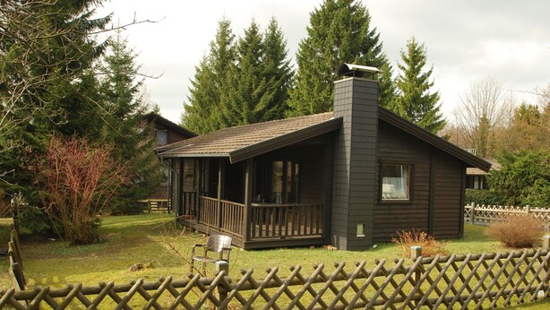 A cabin with a covered porch, surrounded by a wooden fence and tall trees.