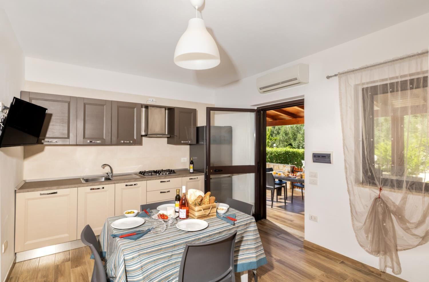 A modern kitchen with a dining area, a flat-screen TV, and an outdoor seating area visible through an open door.