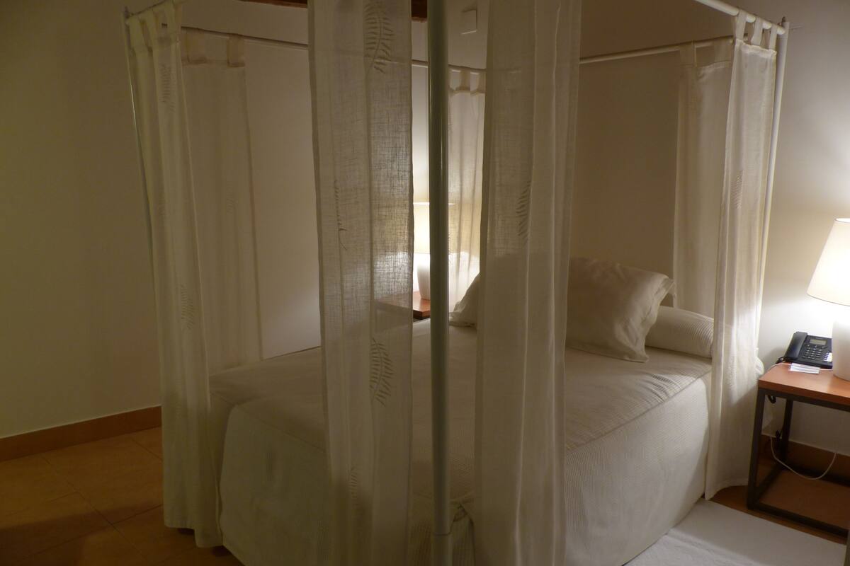 A bedroom with a four-poster canopy bed, a bedside table with a lamp, and a telephone.