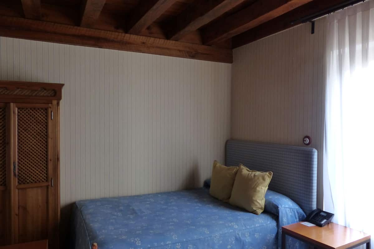 A room with a bed, a wooden wardrobe, a nightstand with a phone, and a window with curtains.