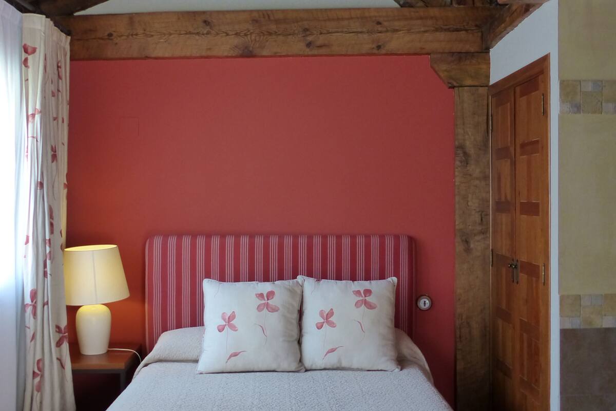 A bedroom with a wooden ceiling, a red wall, a bed with floral pillows, a bedside lamp, and a wooden door.