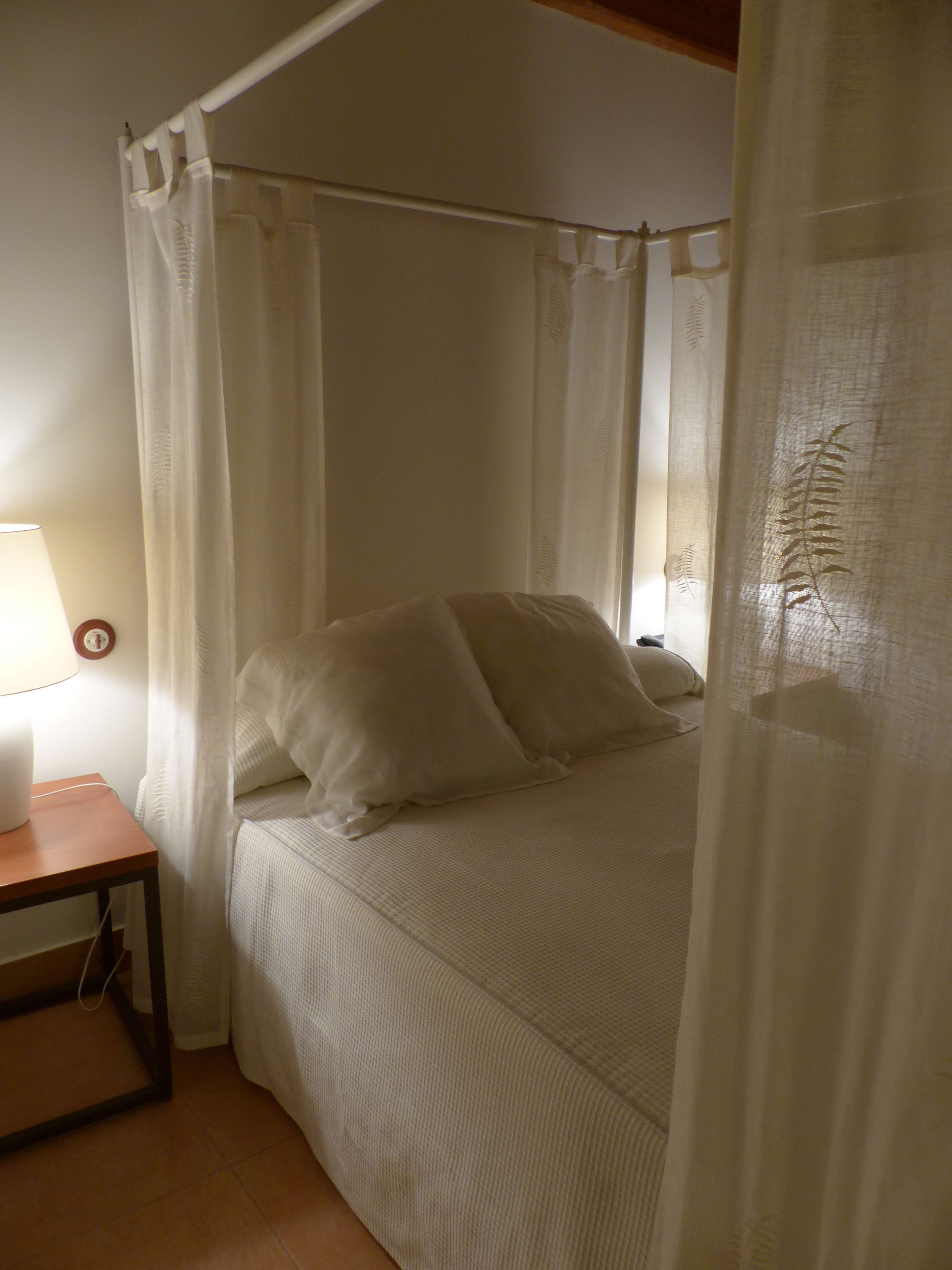 A bedroom with a canopy bed, a bedside table with a lamp, and a window with sheer curtains.