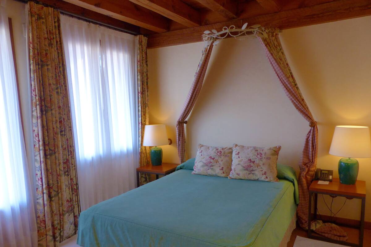 A bedroom with a bed, two bedside tables with lamps, floral curtains, and a wooden ceiling.
