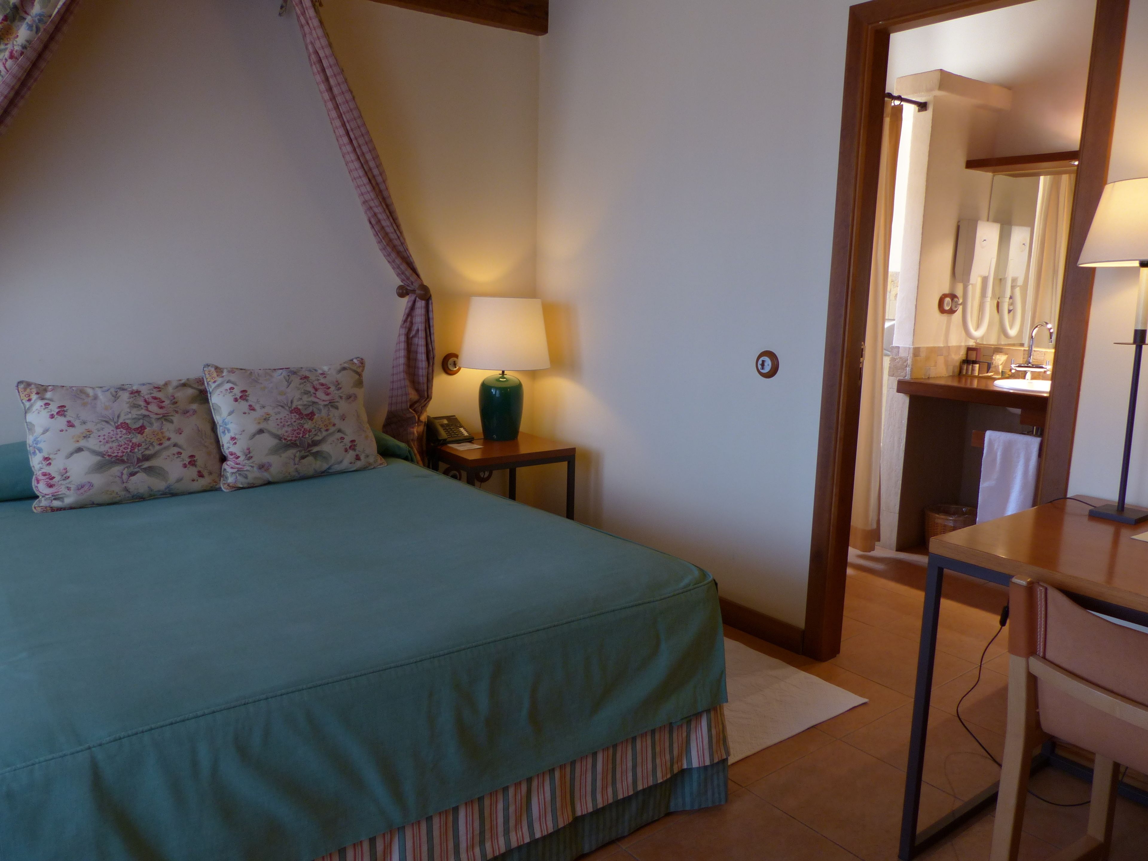 A bedroom with a bed, a bedside table with a lamp, a nightstand, and a bathroom visible through an open door.
