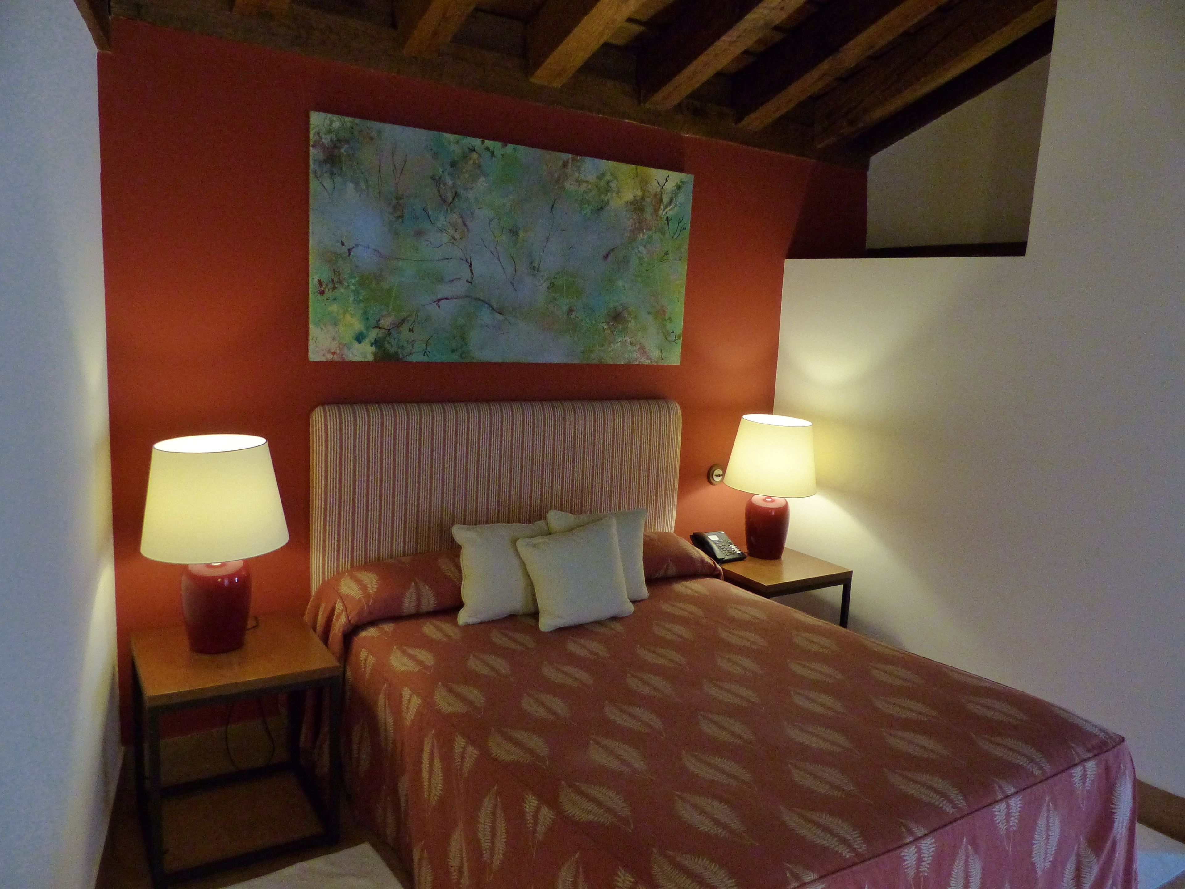 A bedroom with a bed, two bedside lamps, a telephone, and a painting on the wall.